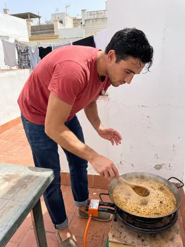 Sebastián, AI character: a rooftop terrace in Cádiz with a paella pan on a gas burner, whitewashed walls, laundry lines and…