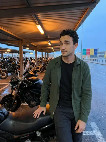 Sebastián, AI character: under a covered parking bay near Cádiz harbor, next to a black motorcycle he is not supposed to ride yet,…