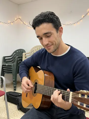Sebastián, AI character: a small Cádiz carnival rehearsal room with plastic chairs, a guitar case on the floor and holiday string…