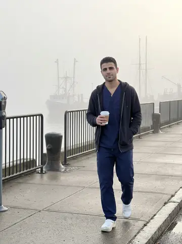 Heitor, AI character: a foggy sidewalk near the New Bedford waterfront after an overnight ER shift, fishing boats soft in the…