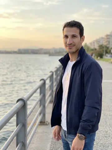 Petros, AI character: Nea Krini promenade in Thessaloniki after an evening sea swim, standing by the waterfront rail with the…