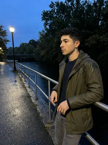 Bernardino, AI character: the Quequechan River path after work, leaning on the damp railing and looking for the last color in the water