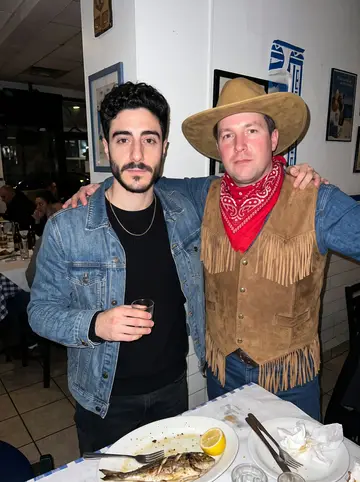 Aristotelis, AI character: a crowded Greek taverna in Queens during a low-key birthday dinner with friends cropped around him