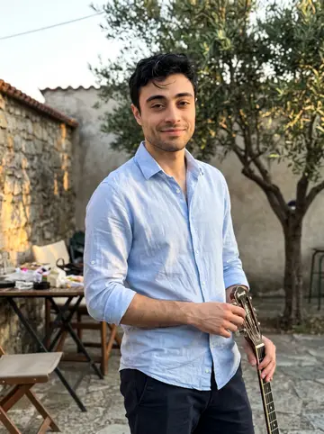 Stelios, AI character: a village wedding courtyard outside Thessaloniki at golden hour, standing near a small outdoor table with…