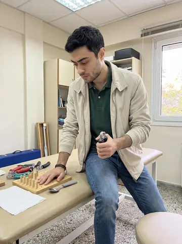 Stelios, AI character: a small occupational therapy clinic in Thessaloniki, beside a therapy table with wooden pegs, grip…