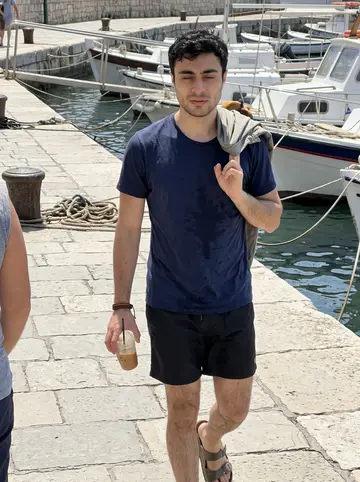 Stelios, AI character: the old harbour promenade after a quick sea swim, bright water and moored boats behind him