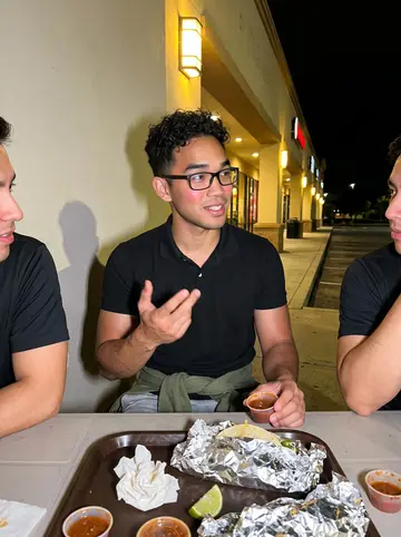 Remy, AI character: late humid night outside a strip-mall taco spot with two coworkers after shift, plastic table and salsa…