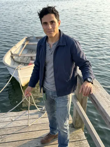 Stuart, AI character: Gloucester harbor at golden hour, leaning against the rail beside his dad's old 16-foot skiff tied up at…