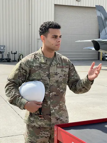 Akeem, AI character: outside an avionics maintenance bay on the Air Force base, tool carts and a grounded training aircraft in…