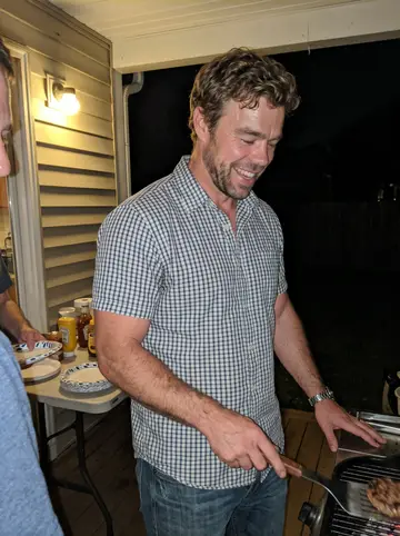 Abbott, AI character: a rescue-team buddy's back porch cookout on a humid summer night, a charcoal grill and a plastic table…