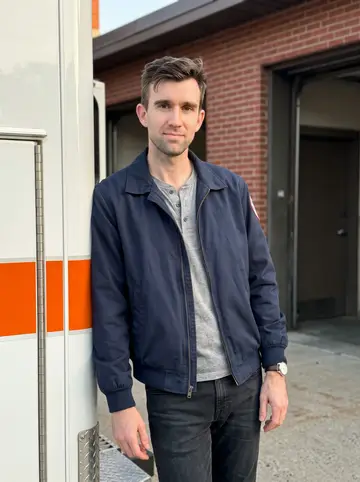 Shawn, AI character: outside the Worcester EMS station bay at the end of an early fall shift, leaning against the open side of…