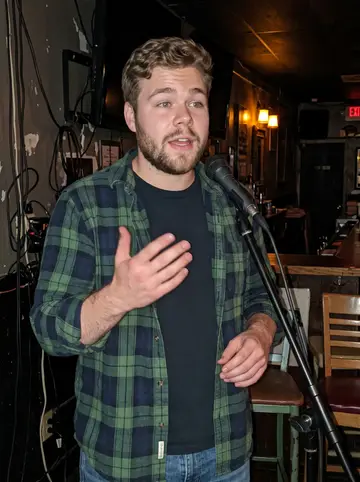Brent, AI character: tiny Pittsburgh open-mic bar with a small microphone stand and a row of mismatched chairs, no readable signs