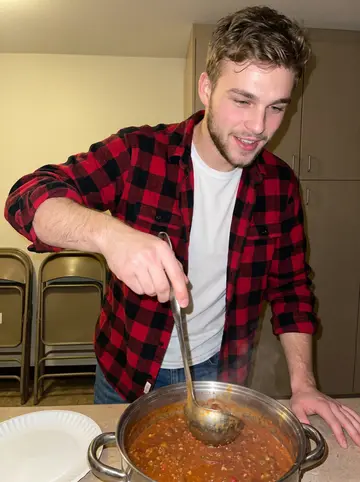 Logan, AI character: the fire hall kitchen during Friday supper, huge chili pot on the counter and folding chairs behind him