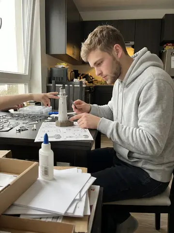 Logan, AI character: the small apartment dining table by the window, model lighthouse kit pieces spread out, his younger…