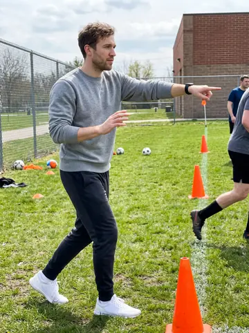 Brian, AI character: a Sunday morning youth soccer field beside a Dayton elementary school, orange cones scattered on damp grass