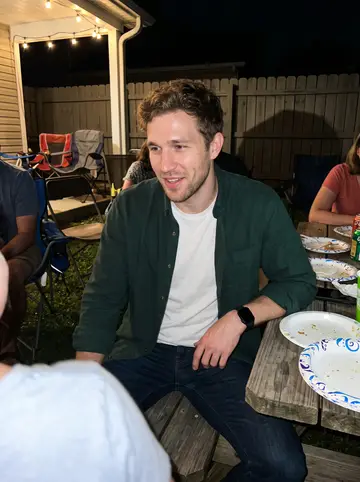 Brian, AI character: his sister's backyard cookout in Dayton, folding chairs and paper plates behind him, relatives cropped at…