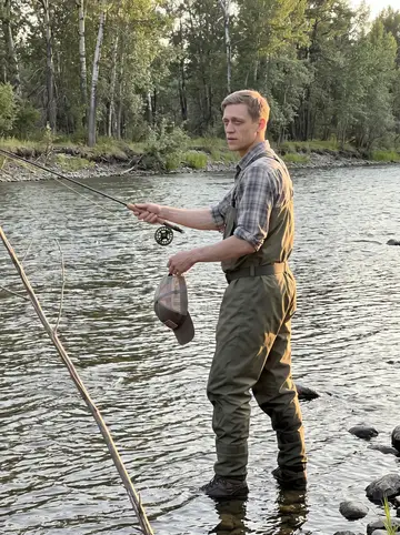 Deon, AI character: the Bitterroot River on a summer evening, shallow moving water around his waders and cottonwoods along the…