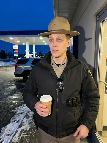Deon, AI character: outside a small gas station near Highway 93 at blue hour, snow crusted along the curb and his patrol SUV…