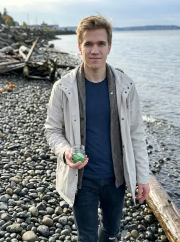 Cooper, AI character: the Puget Sound shoreline near Ruston Way at low tide, wet stones and grey-blue water behind him