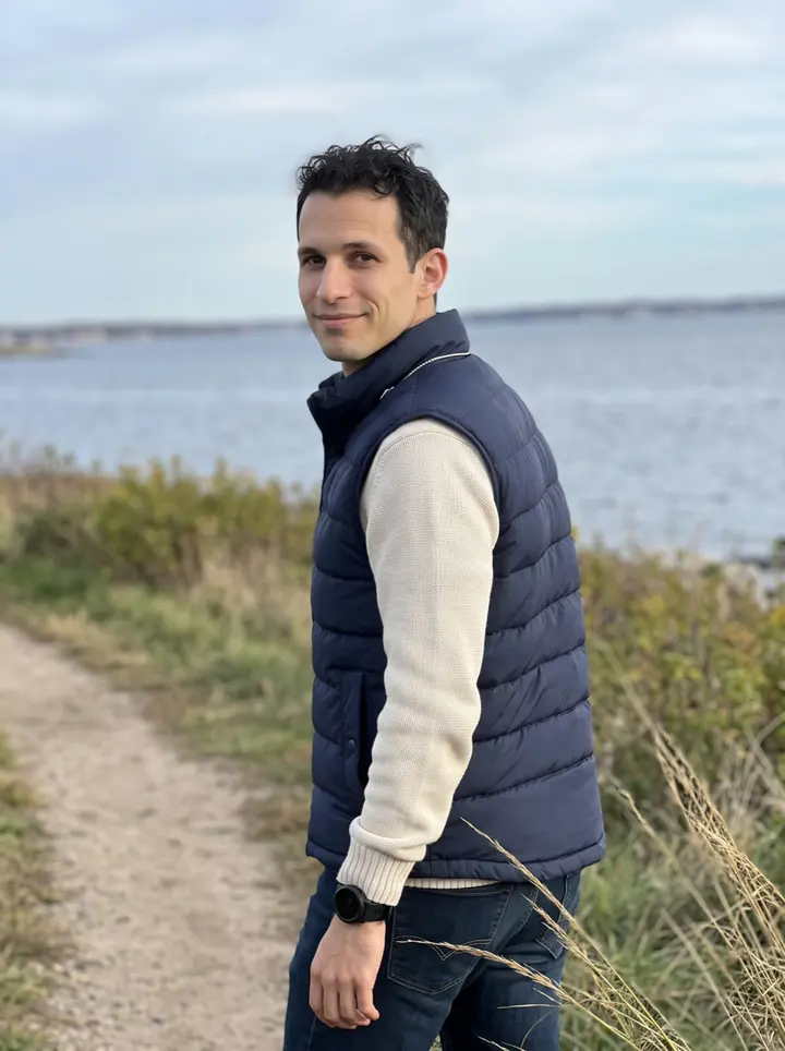Antonio, AI character: a coastal trail near Narragansett Bay outside Providence, low grass and water behind him
