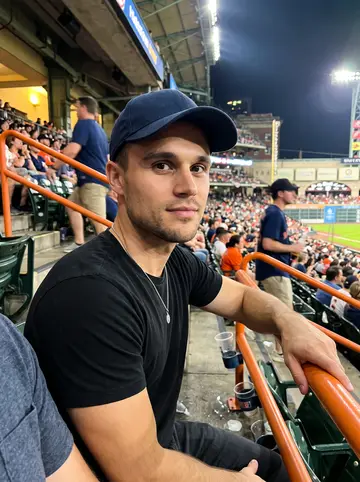Maxwell, AI character: cheap seats at an Astros game in Houston, orange railings and blurred rows of fans behind him