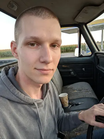 Bram, AI character: the edge of a Route 40 gas station outside Zanesville, standing beside an old dark-blue pickup with the…