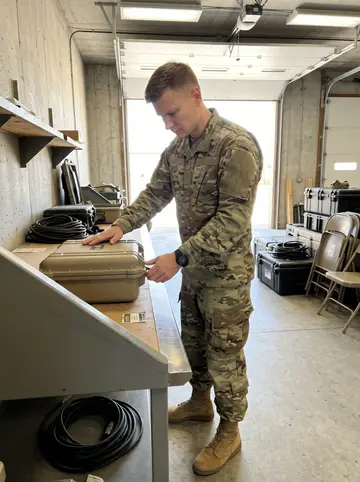 Kalle, AI character: a concrete training bay at Fort Leonard Wood during a daytime equipment check, open bay door behind him…