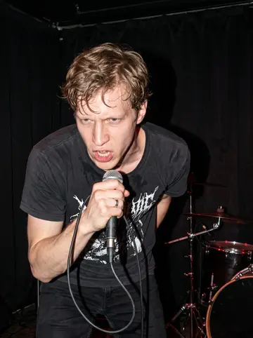 Jiří, AI character: a small Edmonton bar stage during a weekend metal show, drum kit and dark curtain behind him