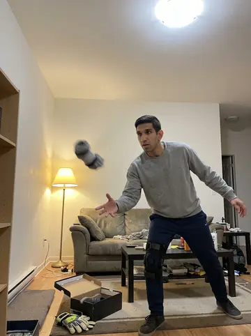 Khaled, AI character: his apartment living room, phone propped on a bookshelf across the room, a rolled-up sock flying through…