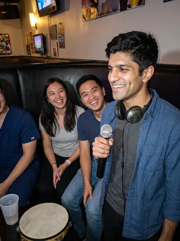 Shreyas, AI character: a small Atlanta karaoke room with his ER shift friends just outside the crop, vinyl booth and colored…