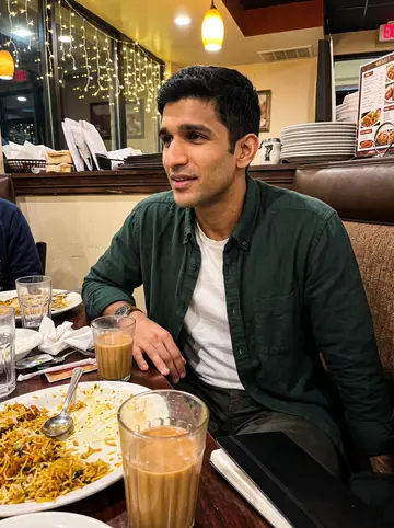 Salman, AI character: a crowded Houston Pakistani restaurant booth during a family dinner, plates of biryani and chai on the…
