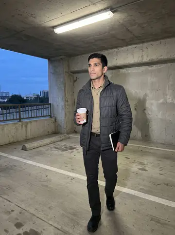 Salman, AI character: a downtown Houston parking garage walkway after work in cool blue evening light, walking toward his car…