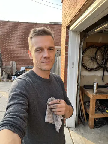 Kacper, AI character: the open doorway of his small Chicago garage off the alley, bicycle wheels hanging behind him and his…
