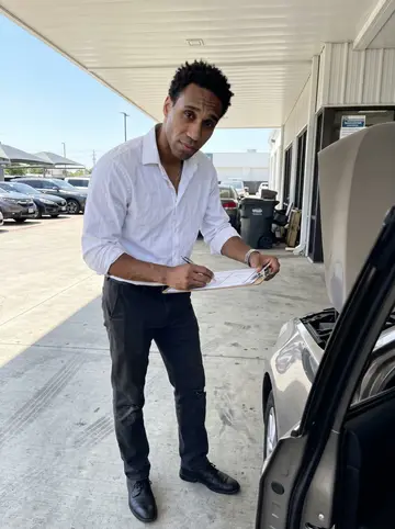 Trevon, AI character: the dealership service lane at midday, between a row of trade-ins and the open door of the sales office