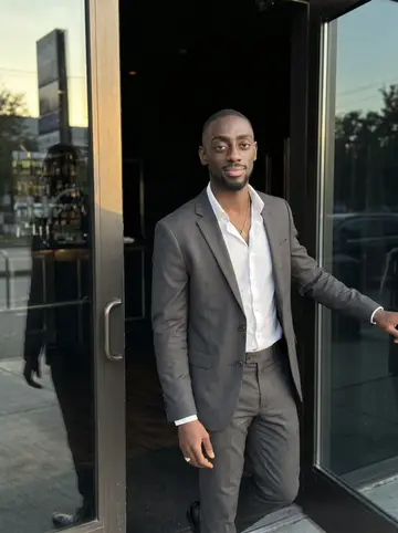Seyi, AI character: outside the dark glass entrance of his Midtown Houston nightclub before opening, one hand resting on the…