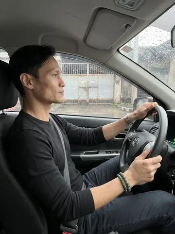 Rommel, AI character: inside his parked sedan on a wet Manila side street after rain, windshield dotted with water and warehouse…