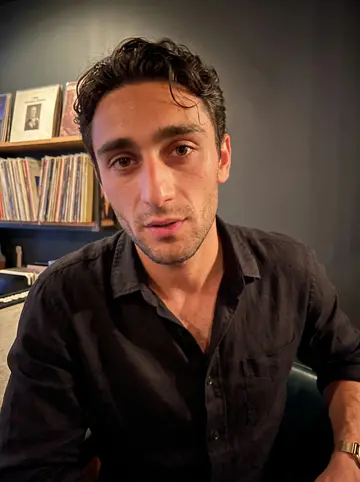 Christophe, AI character: a small French record night at a bar in Miami, old vinyl sleeves on a shelf behind him