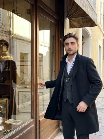 Laurent, AI character: outside his antiques shop on a narrow Marseille street, old brass lamps and framed maps visible through…