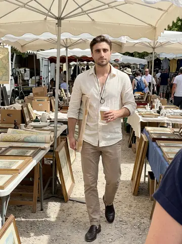 Laurent, AI character: an outdoor antiques market near Provence, folding tables with old frames, maps and ceramic pieces behind him