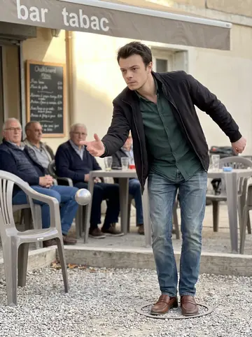 Aurélien, AI character: a pétanque court outside a bar tabac in Marseille, older men watching from plastic chairs beside a…