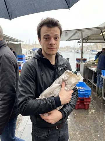 Aurélien, AI character: Marché aux Poissons near the Vieux-Port on a rainy spring morning, wet pavement and fish crates behind him