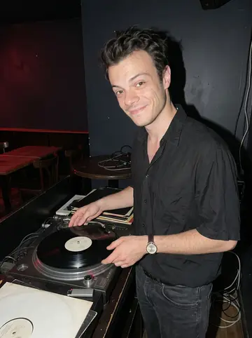 Aurélien, AI character: the small back booth at La Cale after closing, vinyl jazz records stacked beside the turntable and empty…