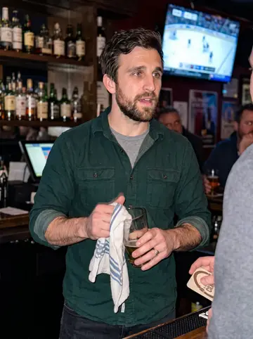 Barry, AI character: behind the bar on a packed winter hockey night, with regulars cropped at the edge of the frame and the…