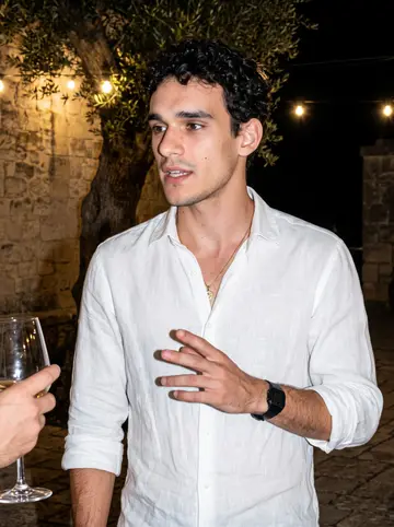 Raffaele, AI character: the stone courtyard of the family masseria late at night during a wedding reception, string bulbs and an…