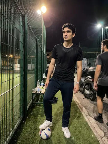 Gennaro, AI character: a fenced five-a-side football pitch in the Quartieri after a Thursday night game, green turf and metal…