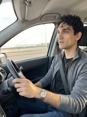 Gennaro, AI character: inside his car on the Lungomare, windows down, Bay of Naples light passing across the dashboard