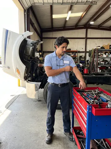 Benicio, AI character: inside his truck repair shop in Laredo, between an open semi hood and a rolling tool cart
