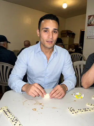Kevin, AI character: a small Jackson Heights social club during Sunday dominoes, plastic table, old men cropped around the edges