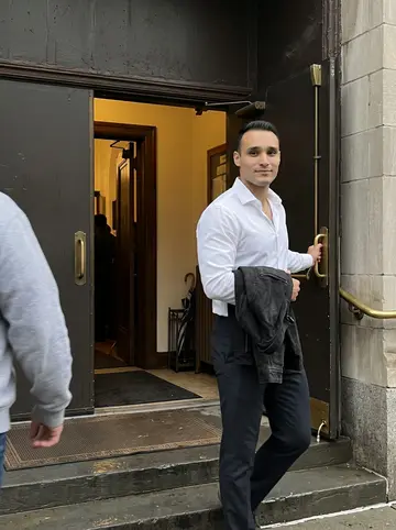 Kevin, AI character: outside a Queens church after rain, holding the side door open while family members move past out of frame