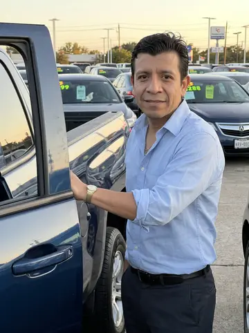 Juan, AI character: the used car dealership lot in Houston near closing time, standing beside a clean dark-blue pickup with…